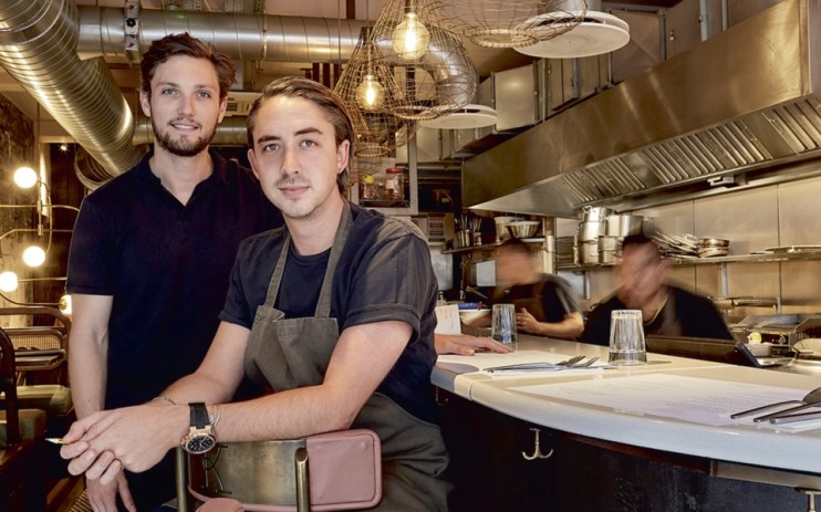 Two men, one in an apron, smiling in a modern Soho restaurant with an open kitchen and industrial decor.