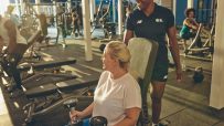 Woman lifting dumbbells with a trainer in a busy gym, promoting fitness and health