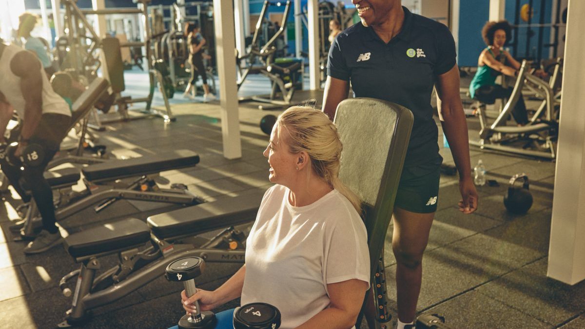 Woman lifting dumbbells with a trainer in a busy gym, promoting fitness and health