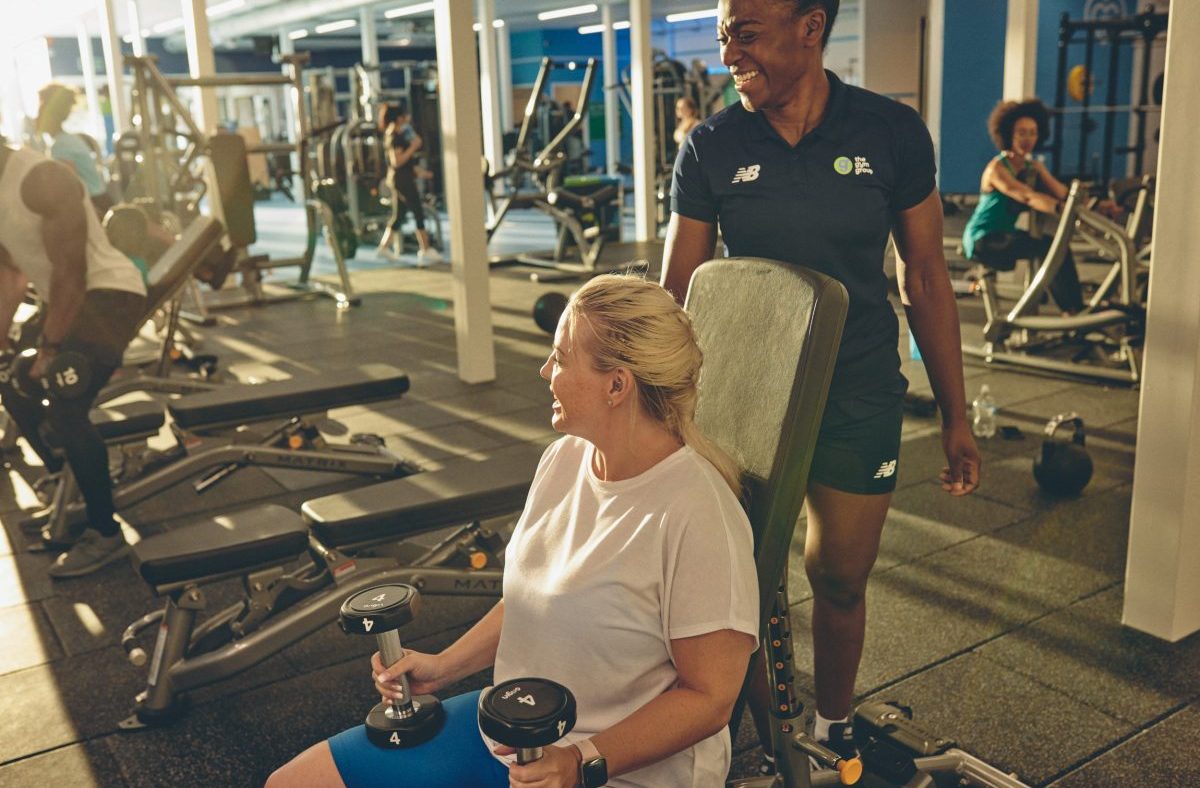 Woman lifting dumbbells with a trainer in a busy gym, promoting fitness and health