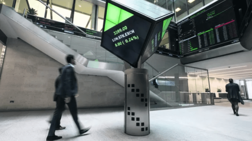 London Stock Exchange interior with a digital display showing LON.STK.EXCH and traders walking past.