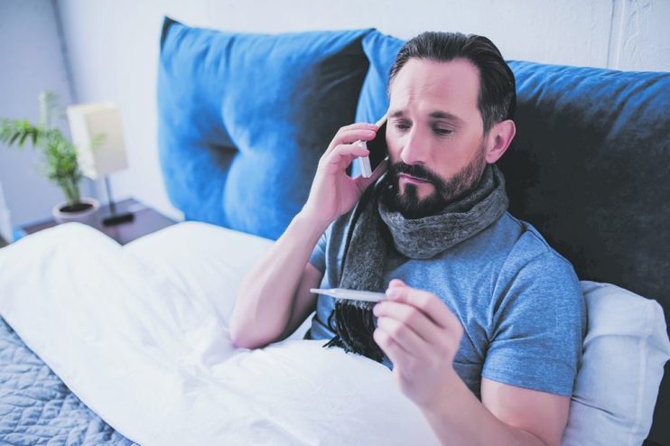 portrait of sick man holding thermometer and calling a doctor while sitting on the bed