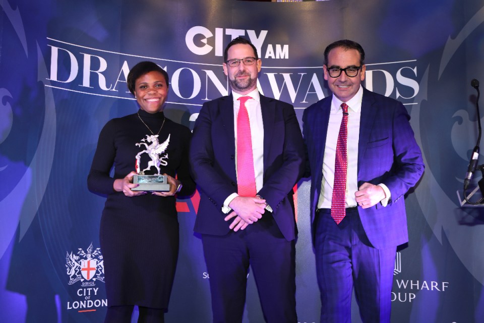 Feyi Osinuga accepts the award on behalf of Janus Henderson's Sarah de Lagarde, alongside Nick Carty of Eastern City BID and awards host Oli Barrett. Morning Wire Dragon Awards 2024 at Mansion House. (Credit: Gretel Ensignia)