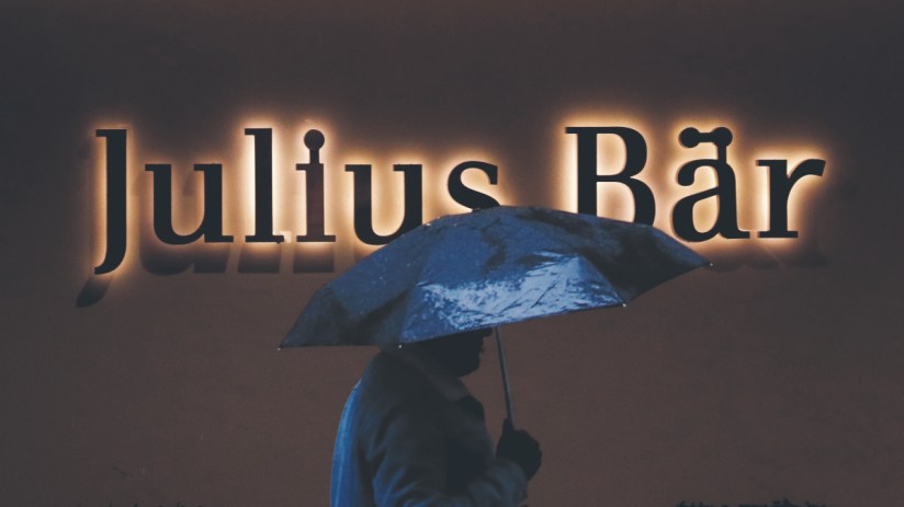 A pedestrian sheltering under an umbrella passes a Julius Baer Group Ltd. branch in Zurich (Photographer: Stefan Wermuth/Bloomberg via Getty Images)