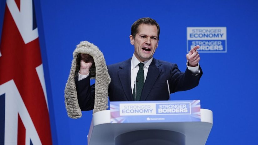 Robert Jenrick speaking at a podium, addressing media about judicial pro-migration bias crackdown, with a serious expression