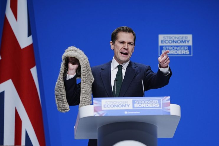 Robert Jenrick speaking at a podium, addressing media about judicial pro-migration bias crackdown, with a serious expression