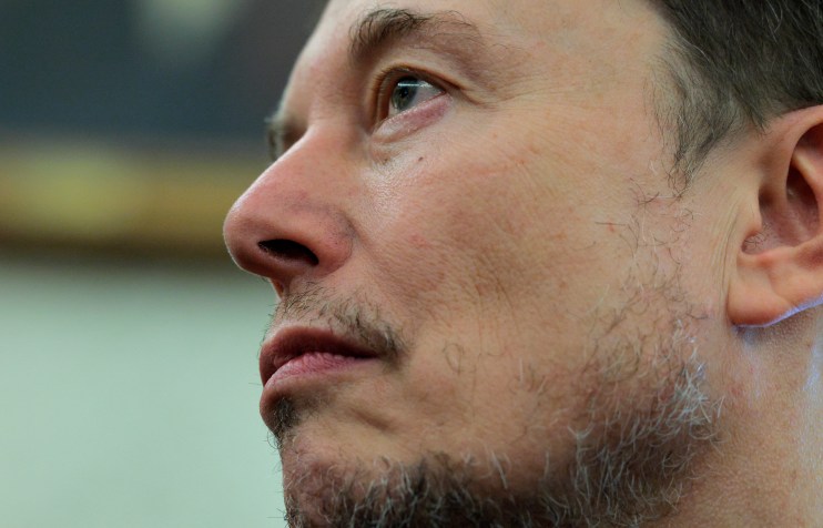 Elon Musk speaking at a business conference, wearing a suit, with a digital backdrop showcasing futuristic technology elem...