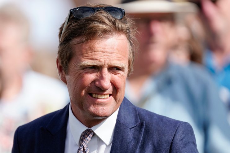 Ralph Beckett speaking at a press conference, wearing a suit, with microphones and reporters visible in the background