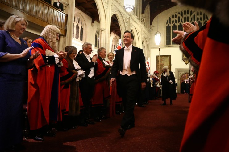 David Cameron speaking at a press conference, wearing a suit and tie, addressing the media with a serious expression.