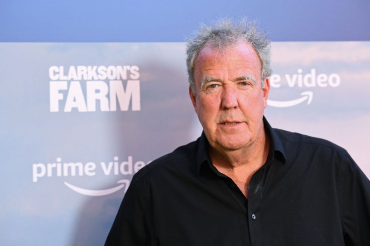 Jeremy Clarkson speaking at a press conference, wearing a suit, gesturing with his hands, addressing a crowd of journalists