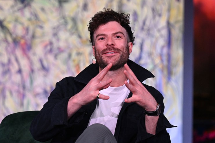 Jordan North in a studio setting, wearing headphones and speaking into a microphone during a radio broadcast.