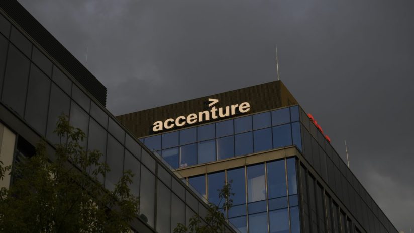 Accenture logo on a glass building facade reflecting a clear blue sky, representing corporate presence and digital innovat...