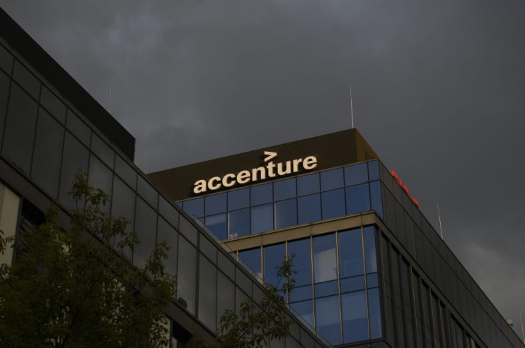 Accenture logo on a glass building facade reflecting a clear blue sky, representing corporate presence and digital innovat...