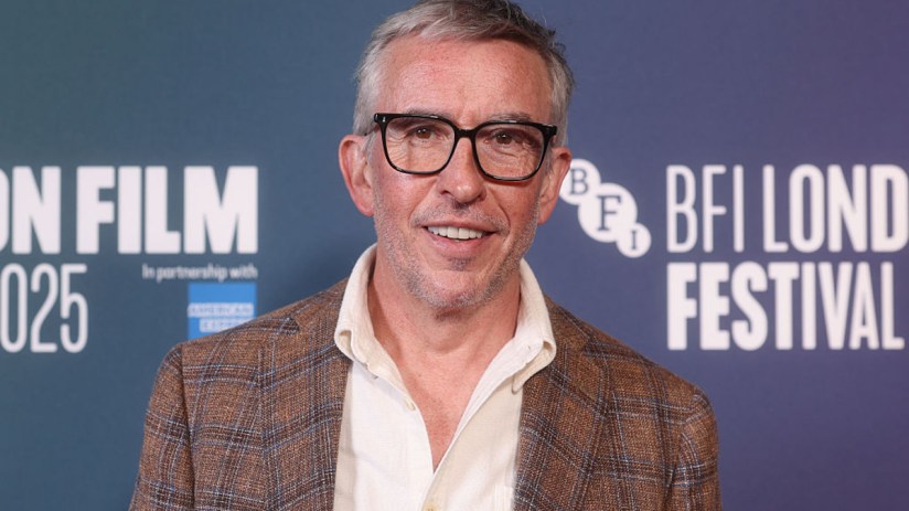 Steve Coogan speaking at a press conference, wearing a suit and tie, addressing the media with a serious expression, micro...