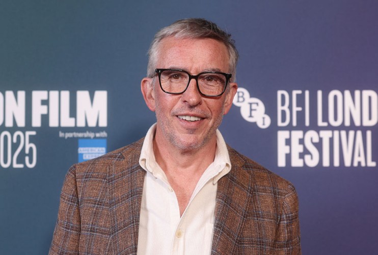 Steve Coogan speaking at a press conference, wearing a suit and tie, addressing the media with a serious expression, micro...