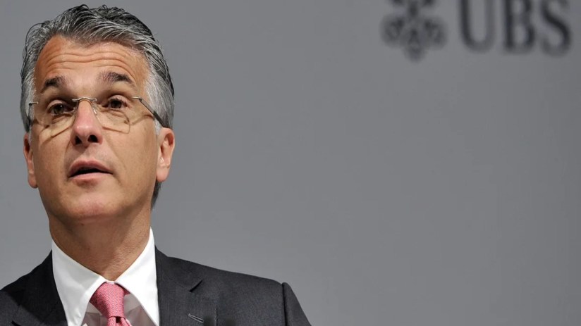 Sergio Ermotti, UBS CEO, addressing media at a press conference, wearing a suit and glasses, with a serious expression