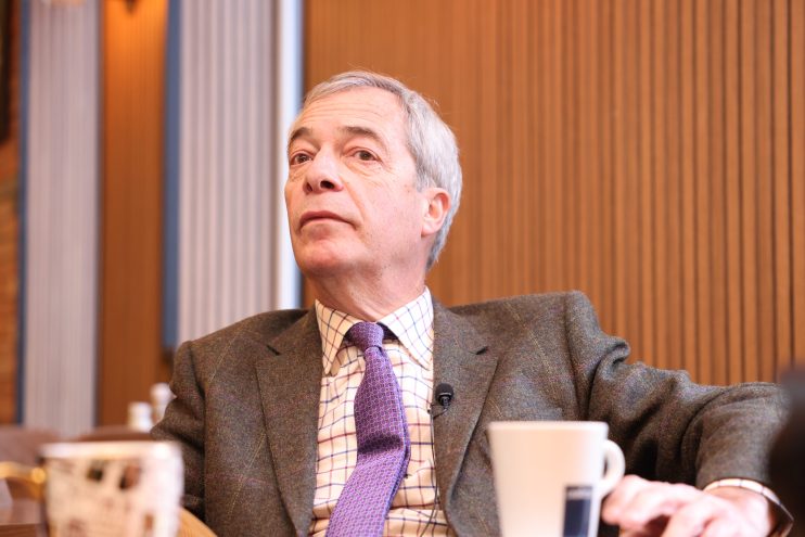 Nigel Farage speaking at a conference podium with audience in background, wearing a suit and tie, emphasizing political di...
