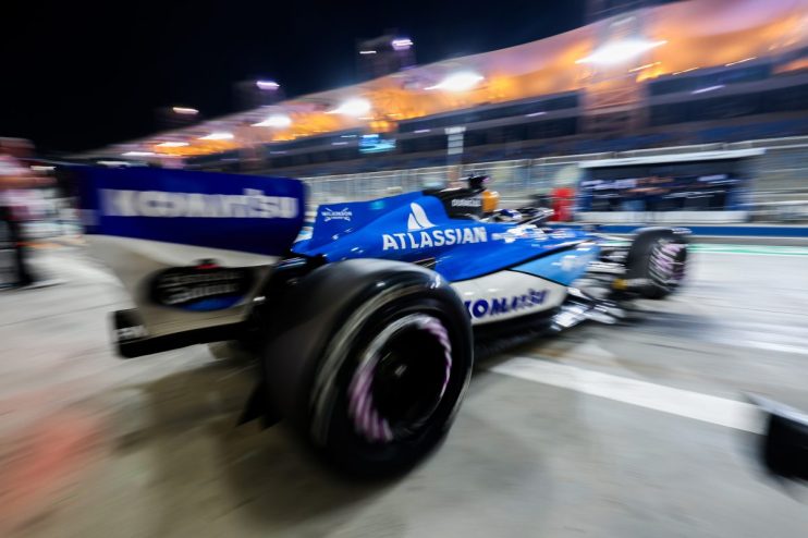 Alex Albon exits Formula 1 garage, wearing racing suit, surrounded by team members at busy pit lane during race weekend.