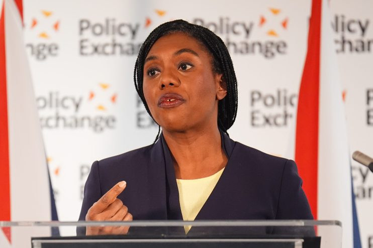 Kemi Badenoch speaking at a podium during a press conference, wearing a navy blazer and addressing the audience confidently