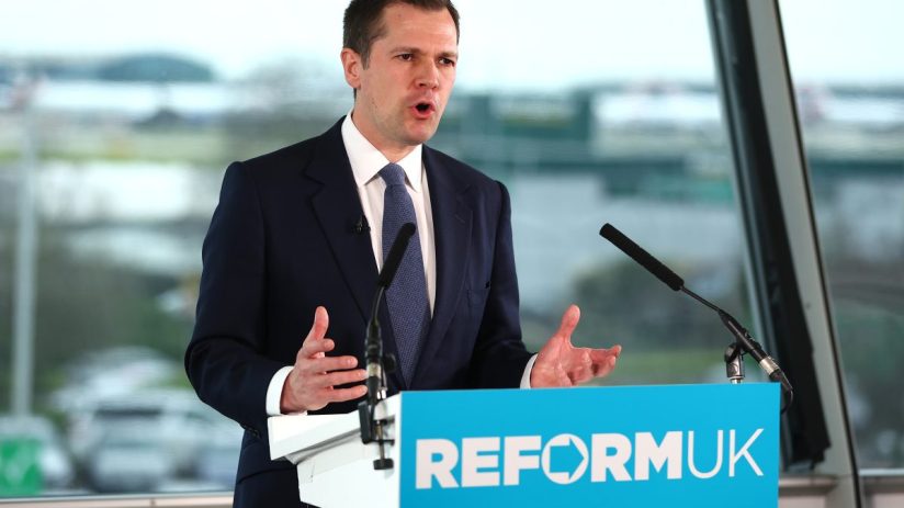 Robert Jenrick speaking at a press conference, addressing current policy issues, wearing a suit and standing behind a podium