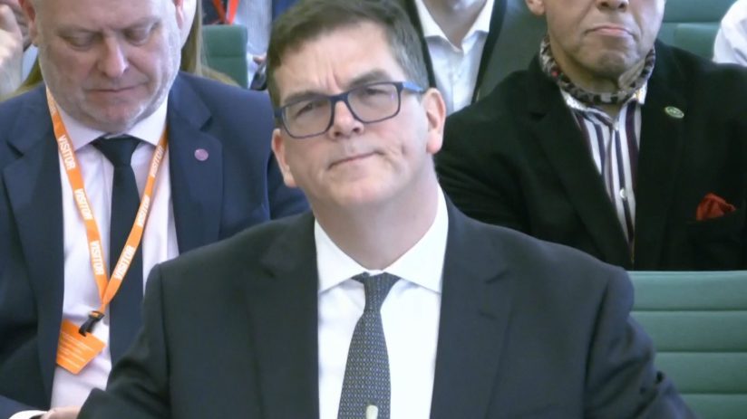 Sir Olly Robbins in a formal setting, possibly during a business or political event, wearing a suit and tie, engaging in d...