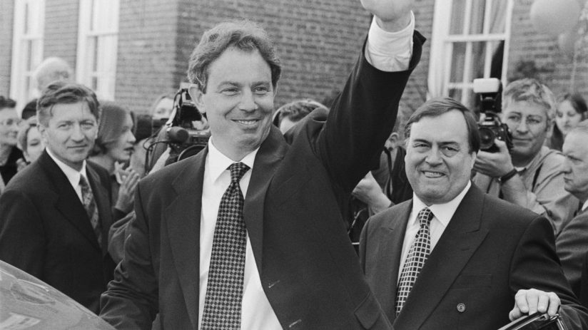 Tony Blair delivering a speech in a formal setting, wearing a suit and tie, with a serious expression, addressing an audience