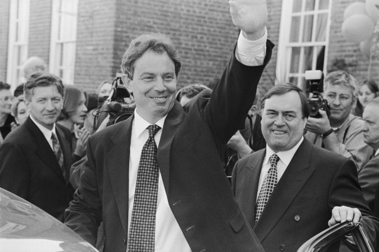 Tony Blair delivering a speech in a formal setting, wearing a suit and tie, with a serious expression, addressing an audience