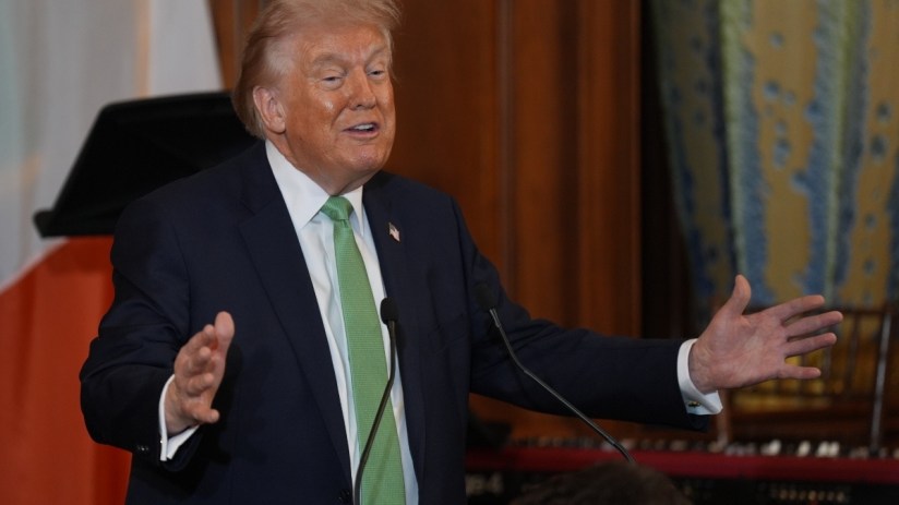 Donald Trump wearing a green tie at a public event, addressing the audience with a serious expression in a formal setting