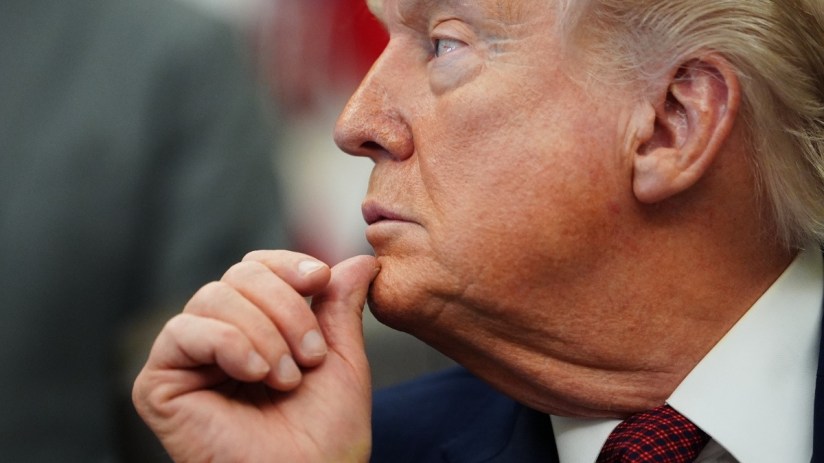 Donald Trump with hand on chin, appearing contemplative during a public event, wearing a suit and red tie.