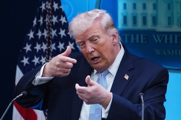 Donald Trump speaking at press conference podium, addressing media with serious expression, American flags in background