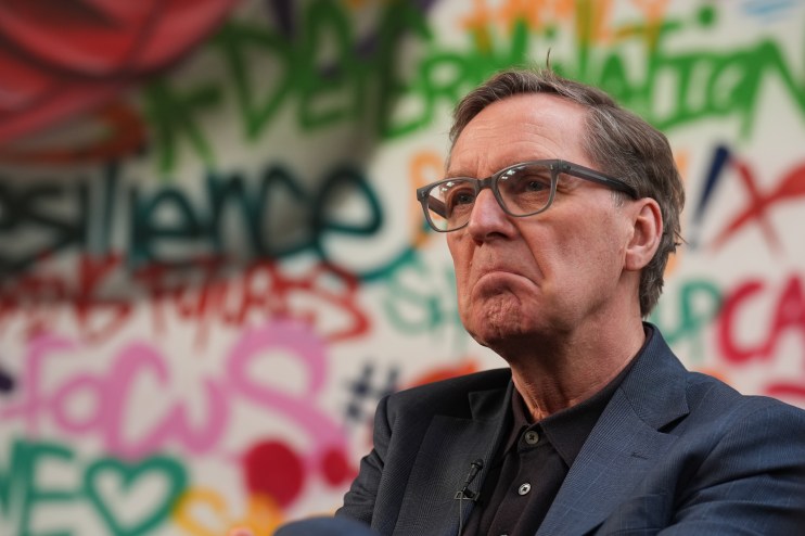 Alan Milburn speaking at a business conference, wearing a suit and tie, discussing economic strategies and policies