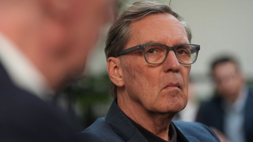 Alan Milburn delivering a speech at a press conference, wearing a suit and tie, addressing economic policy issues.