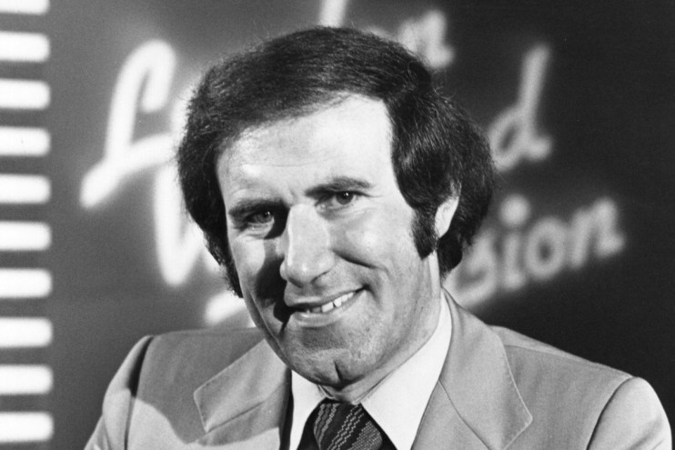 Andrew Neil delivering a speech at a business summit, wearing a suit and tie, with a presentation screen in the background
