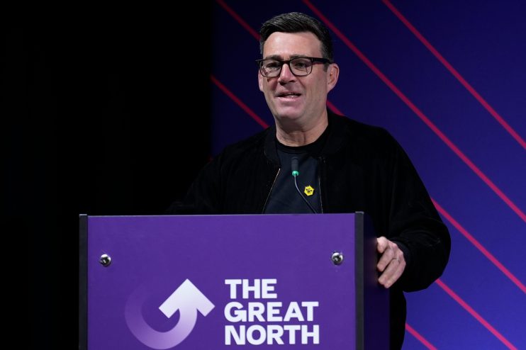 Andy Burnham delivers speech, standing at podium, emphasizing key points, wearing a suit, addressing an engaged audience