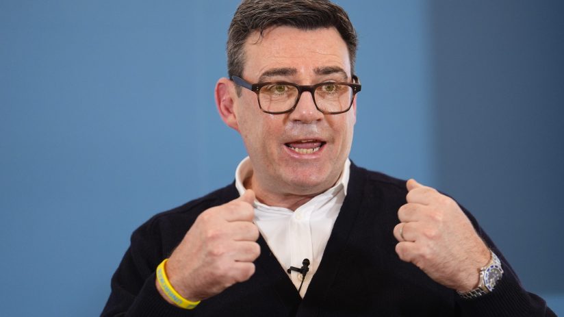 Andy Burnham speaking at a public event, wearing a suit, addressing an audience with a focused expression and engaged deme...