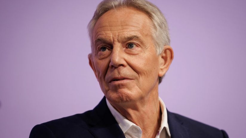 Tony Blair delivering a speech at a conference podium, discussing current global political issues.