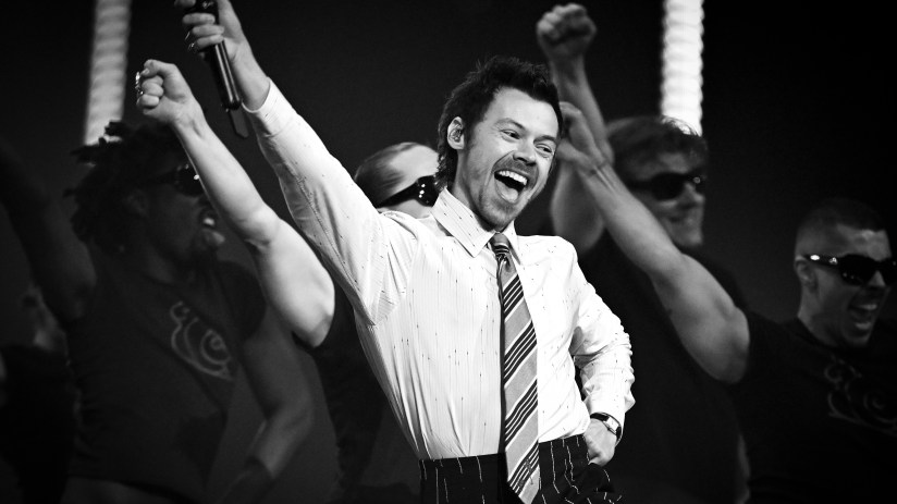 Harry Stykes attending a public event, dressed in a stylish suit, addressing an audience, engaging with fans and media.
