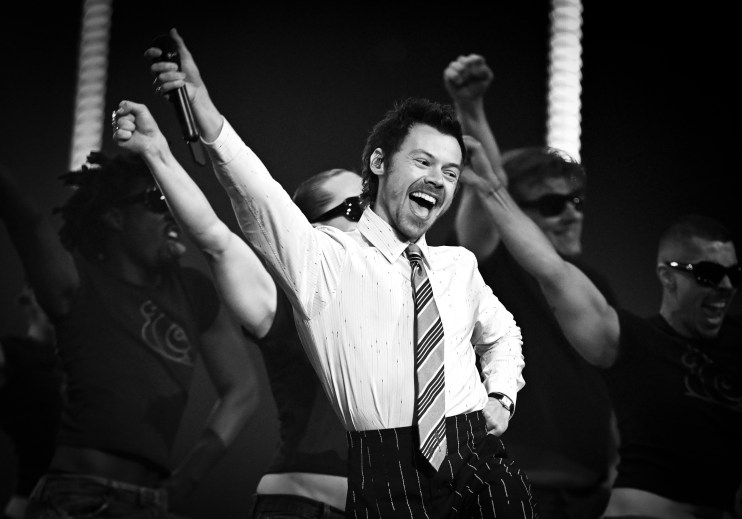 Harry Stykes attending a public event, dressed in a stylish suit, addressing an audience, engaging with fans and media.