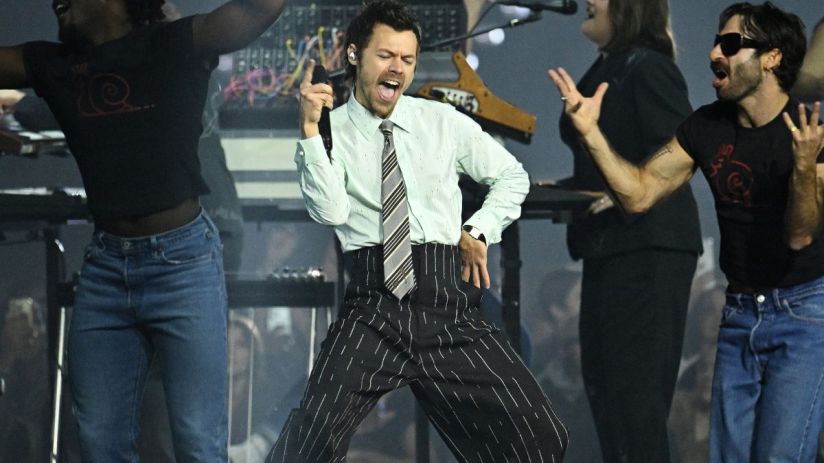 Harry Styles performing on stage with vibrant lighting at a concert, wearing a stylish outfit and engaging with the audience.