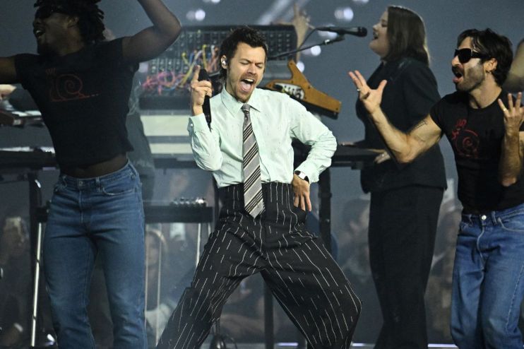 Harry Styles performing on stage with vibrant lighting at a concert, wearing a stylish outfit and engaging with the audience.