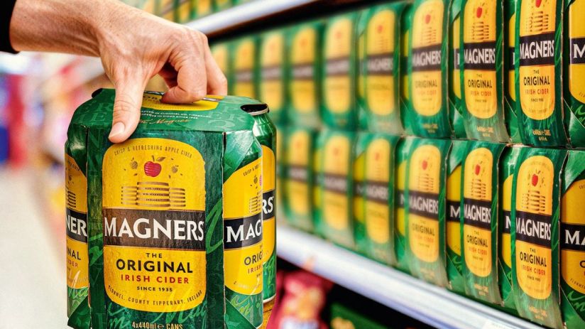 Magners cider bottles displayed on a wooden table with fresh apples and a scenic orchard in the background.