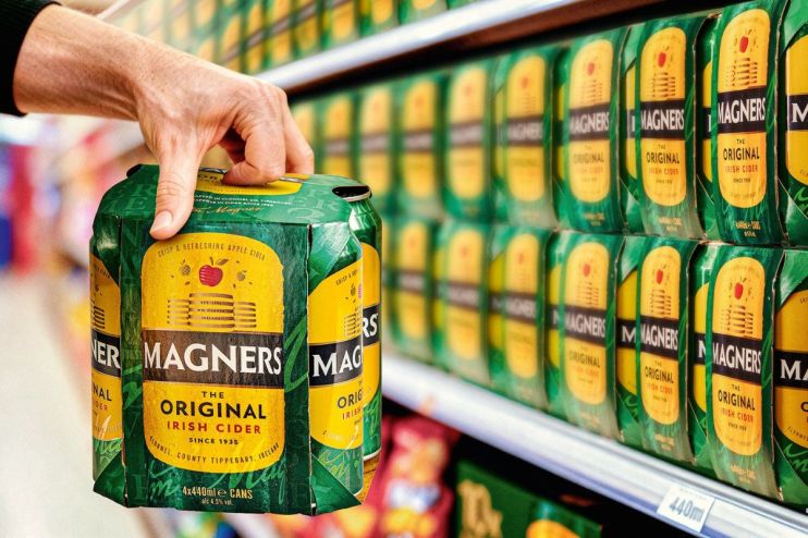 Magners cider bottles displayed on a wooden table with fresh apples and a scenic orchard in the background.