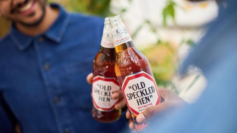 Old Speckled Hen beer bottle with label visible on a wooden table, showcasing its classic design and distinctive branding