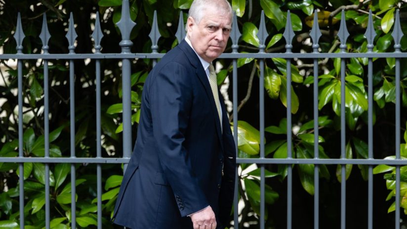 Prince Andrew attending a formal event dressed in a dark suit, engaging in conversation with other attendees in a hall.