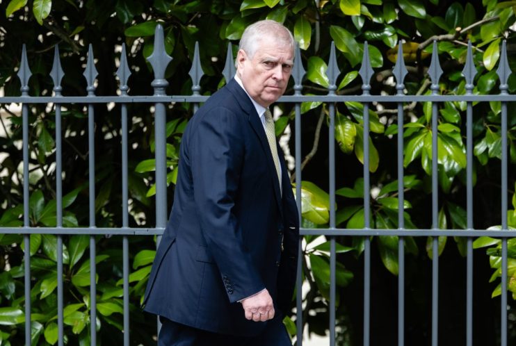 Prince Andrew attending a formal event dressed in a dark suit, engaging in conversation with other attendees in a hall.