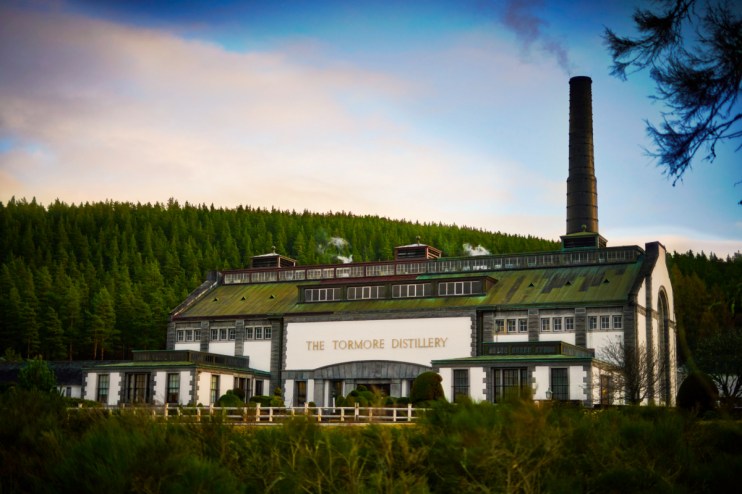 Tormore Distillery in scenic Speyside, showcasing traditional architecture with whisky barrels and lush greenery in the fo...