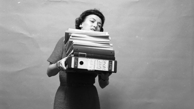1955 secretary overwhelmed by towering stack of files, symbolizing challenges in office management and document handling
