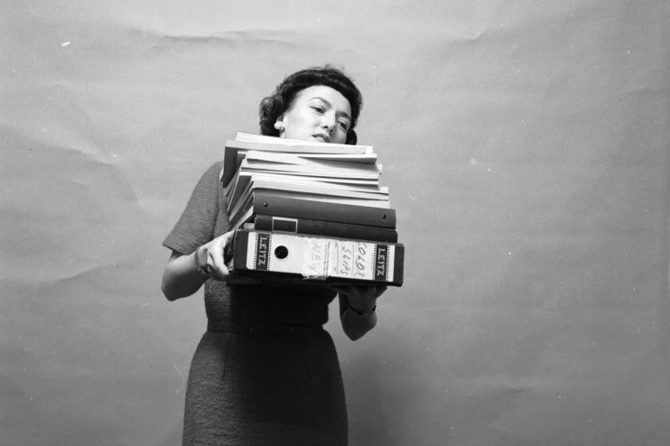 1955 secretary overwhelmed by towering stack of files, symbolizing challenges in office management and document handling