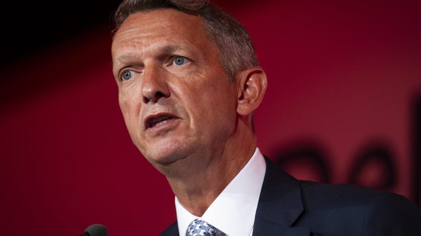 Andy Haldane speaking at a business conference, gesturing with hands, wearing a suit and tie, addressing economic issues.