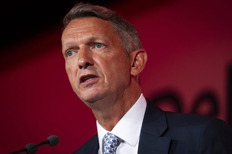 Andy Haldane speaking at a business conference, gesturing with hands, wearing a suit and tie, addressing economic issues.